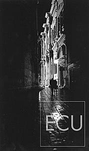 Black and white photo of a couple walking in the rain approaching Campo San Rocco at night in San Polo sestiere of Venice, Italy